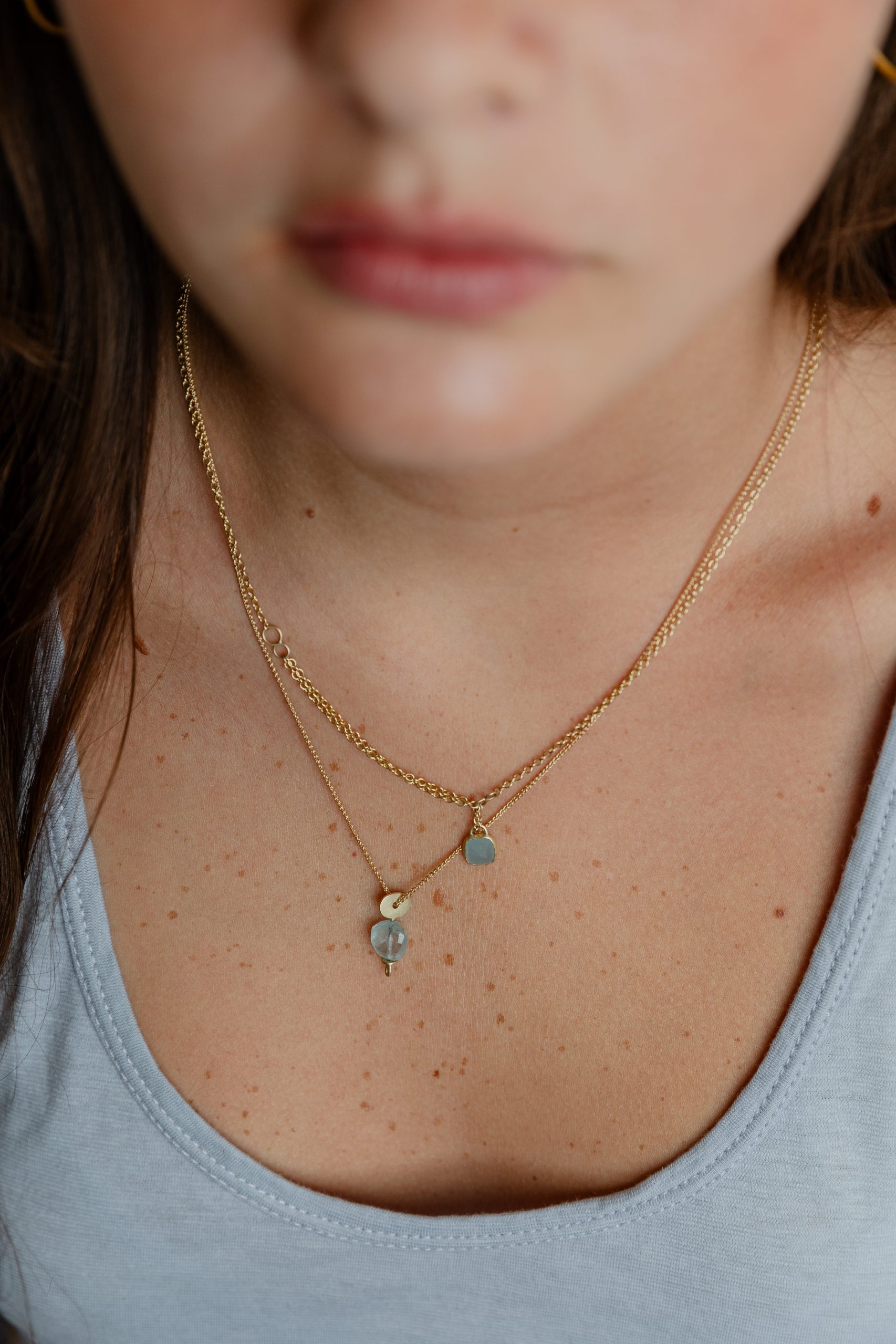 A close-up of a woman wearing two gold necklaces with small pendants, one heart-shaped and one circular, against her light blue top. Her lower face and collarbone are visible.