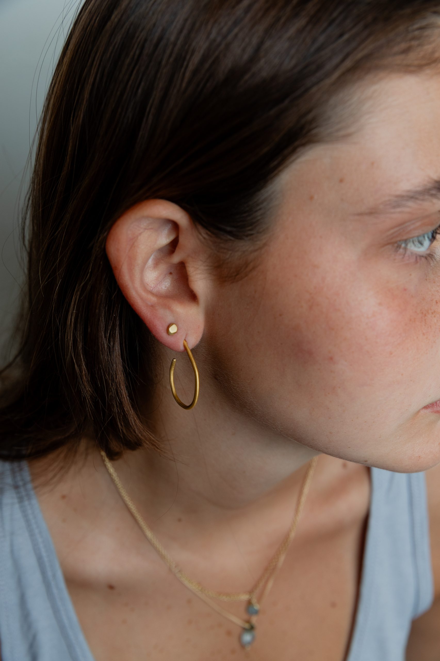 A close-up of a person with light brown hair wearing a gold hoop earring, a small gold stud, and two layered gold necklaces. The person is wearing a gray top and looking slightly to the right.