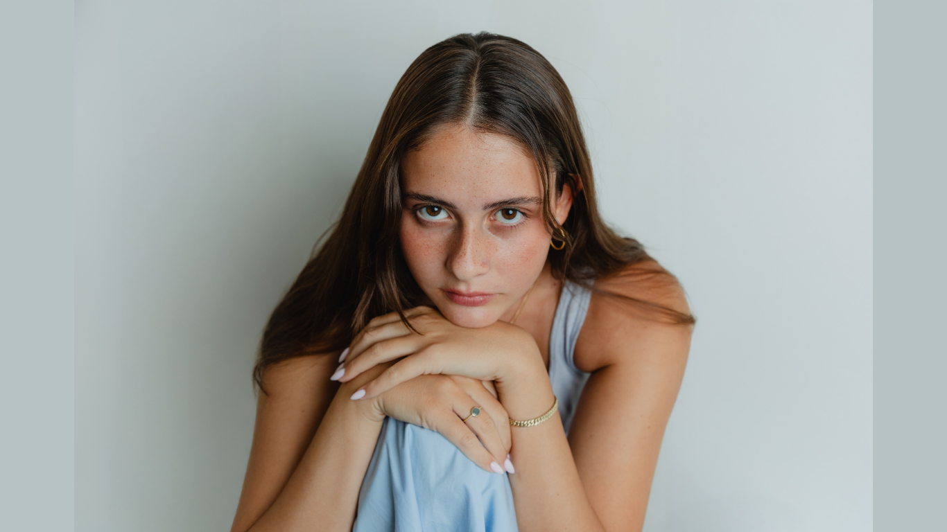 A young woman with long brown hair and freckles sits against a plain light background, resting her chin on her folded hands and looking directly at the camera with a serious expression. She wears a light blue sleeveless top.