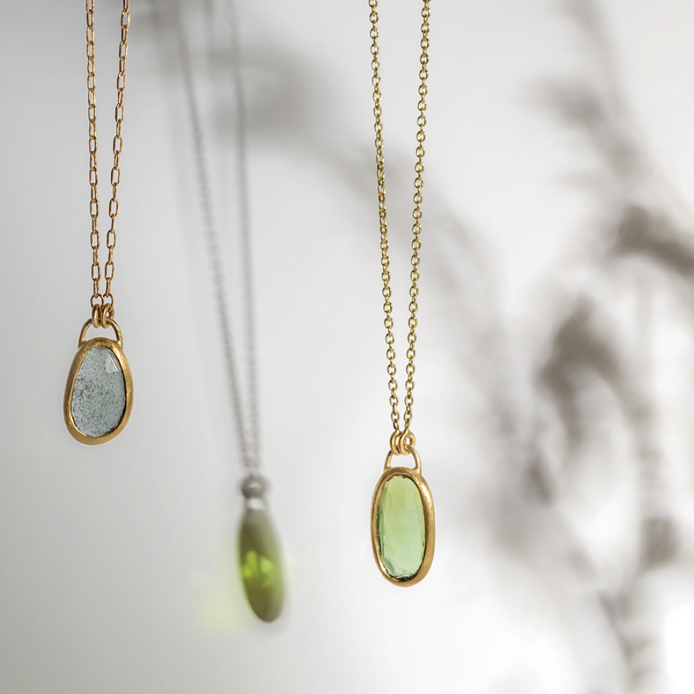 Two gold chain necklaces with oval gemstone pendants—one gray and one light green—hang against a pale, blurred background with soft shadows and reflections.