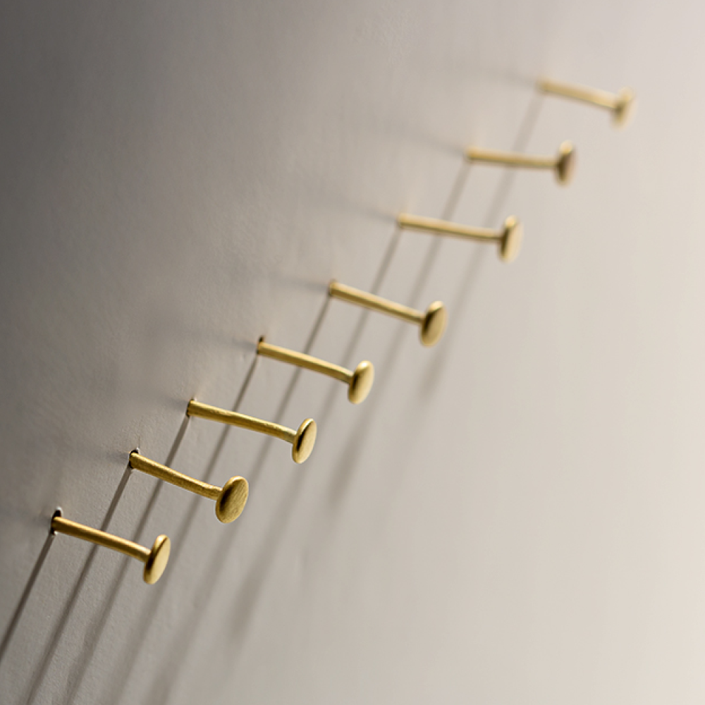 Seven gold nails are partially hammered into a light-colored wall in a diagonal line, casting long shadows to the right. The nails create a pattern of parallel lines with their shadows.