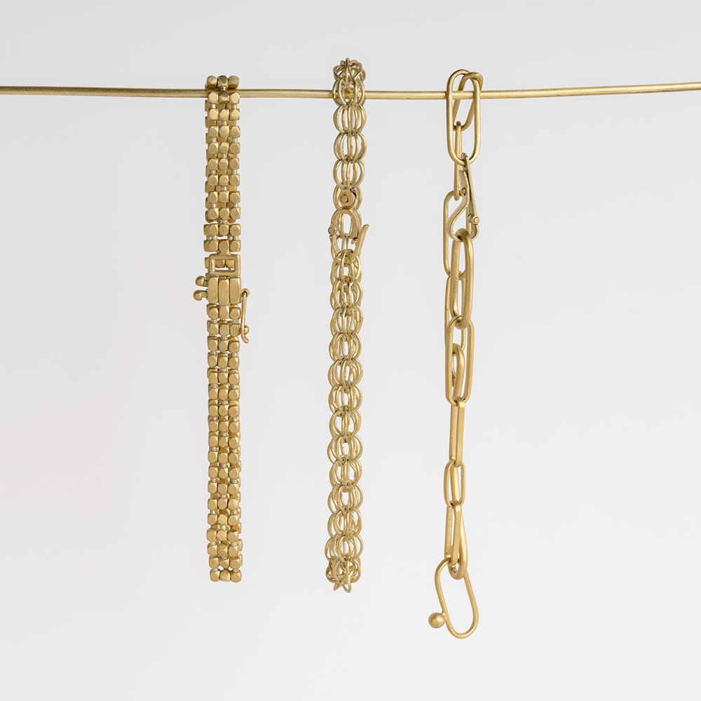 Three gold bracelets with different link designs hang side by side on a thin gold rod against a plain white background.