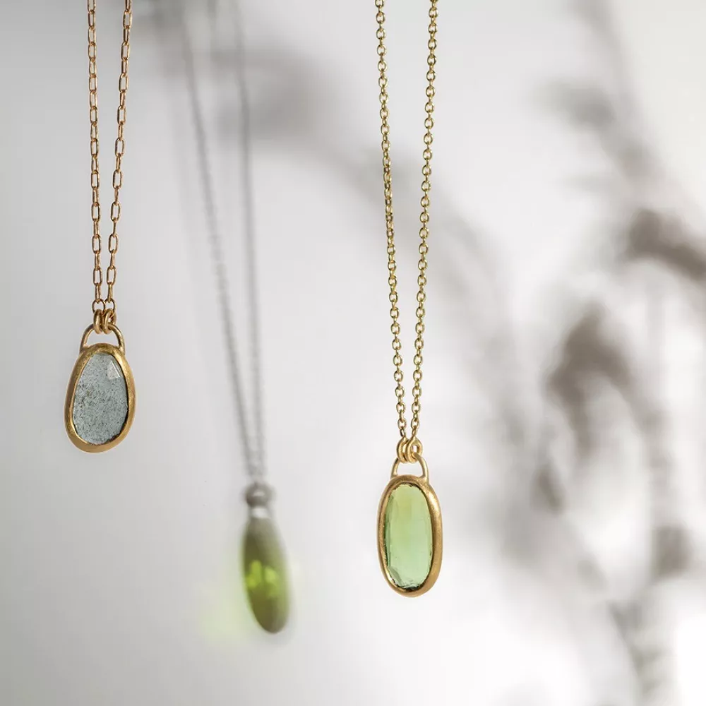 Two gold necklaces hang side by side, each with an oval gemstone pendant—one grayish-blue, one light green—casting soft shadows and colorful reflections on a light background.