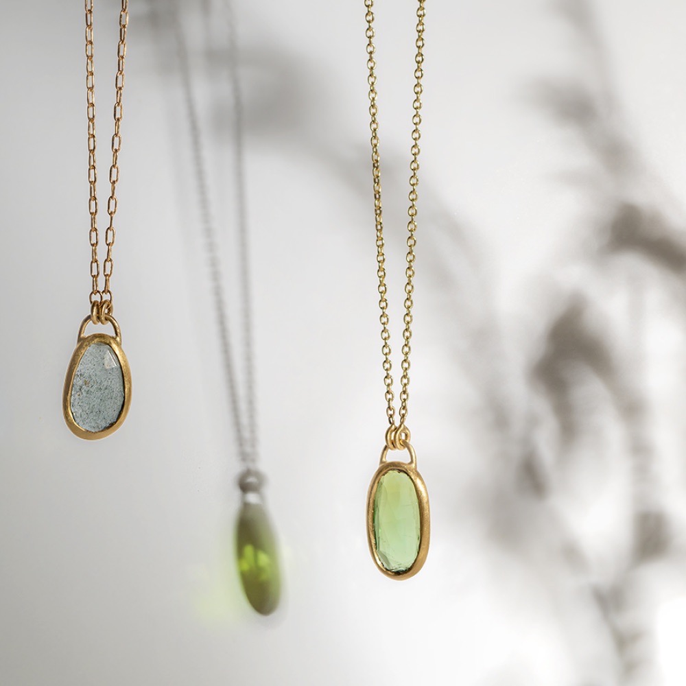 Two gold necklaces hang side by side, each with an oval gemstone pendant—one grayish-blue, one light green—casting soft shadows and colorful reflections on a light background.