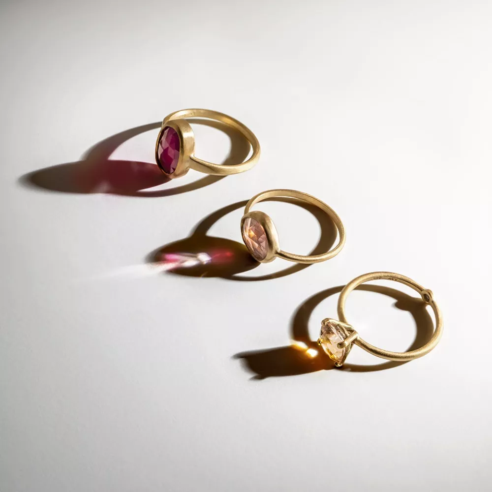 Three gold rings with oval gemstones—one red, one pink, and one clear—are arranged in a row on a white surface, casting colorful light reflections and soft shadows.