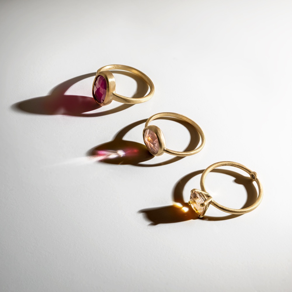 Three gold rings with oval gemstones—one red, one pink, and one clear—are arranged in a row on a white surface, casting colorful light reflections and soft shadows.
