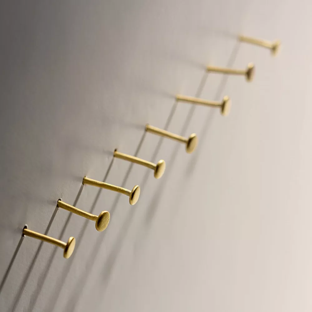 Nine gold nails are arranged in parallel rows on a light-colored wall, casting long shadows to the right, creating a pattern of lines and circles.