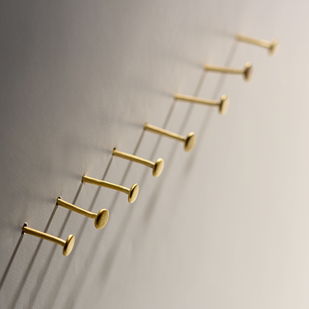 Nine gold nails are arranged in parallel rows on a light-colored wall, casting long shadows to the right, creating a pattern of lines and circles.