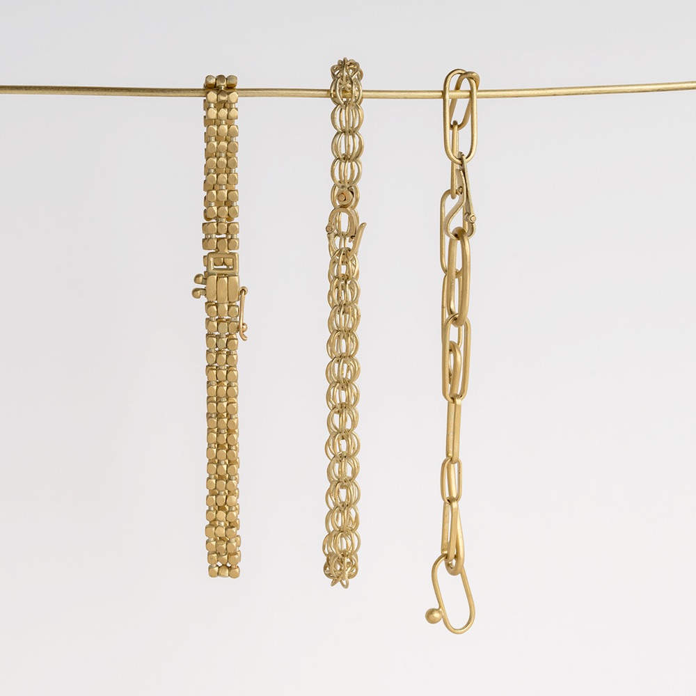 Three gold bracelets, each with a different chain design, hang from a horizontal rod against a plain white background. The left bracelet has small beads, the middle one circular links, and the right one large rectangular links.