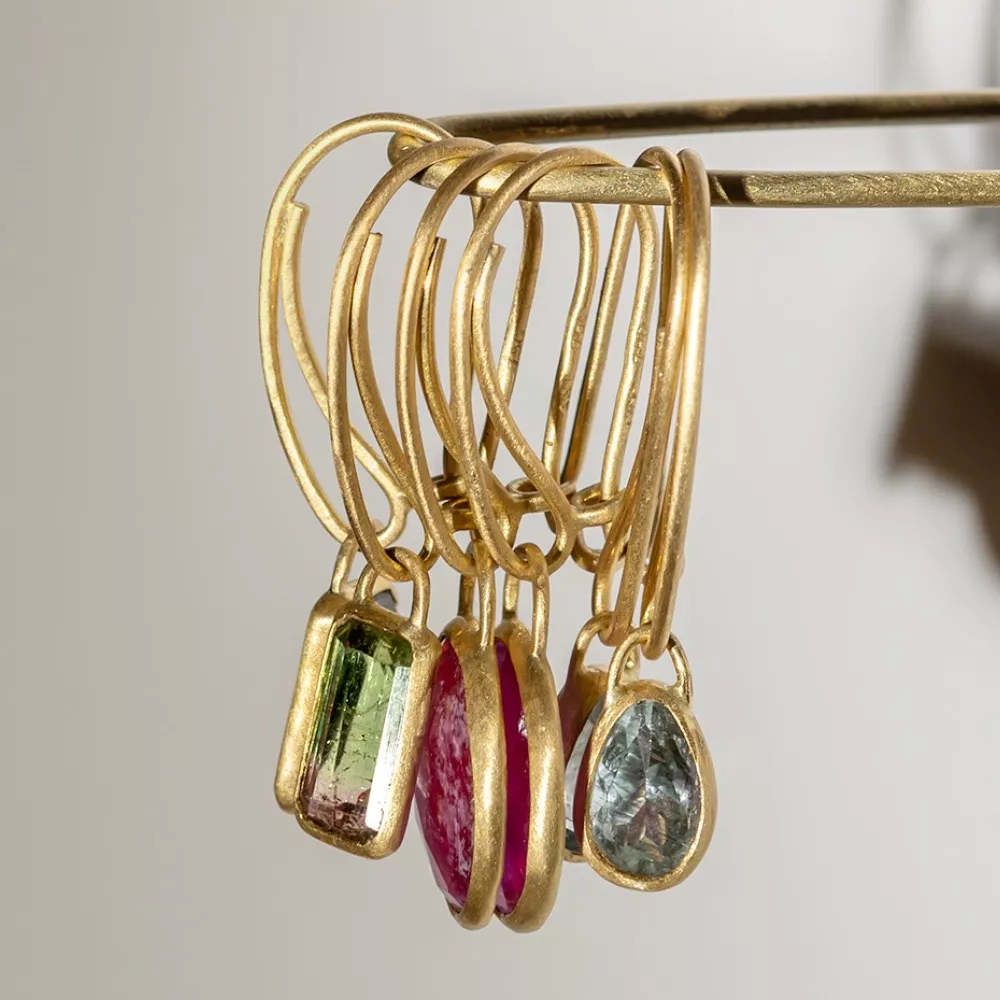 Several gold hoop earrings with colorful gemstone pendants—green, pink, and light blue—are displayed hanging from a horizontal rod against a plain background.