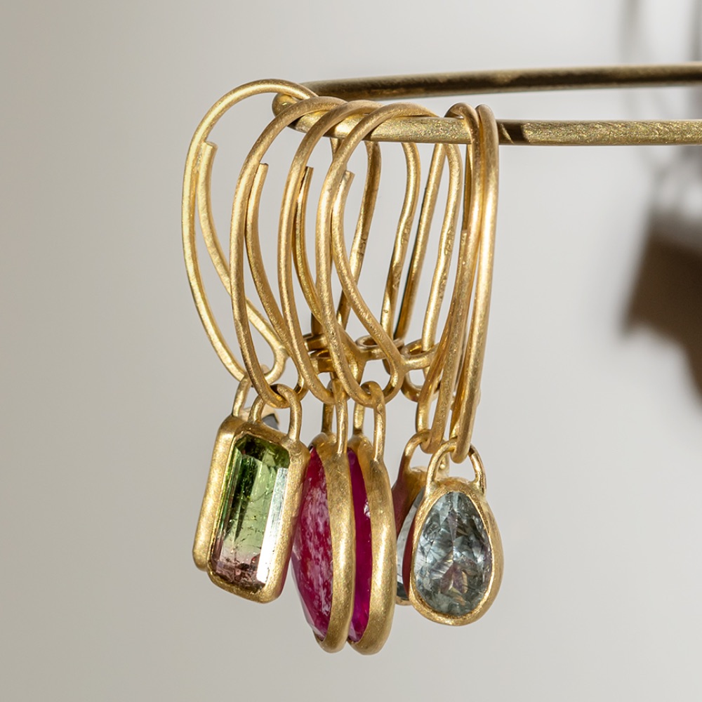 Several gold hoop earrings with colorful gemstone pendants—green, pink, and light blue—are displayed hanging from a horizontal rod against a plain background.
