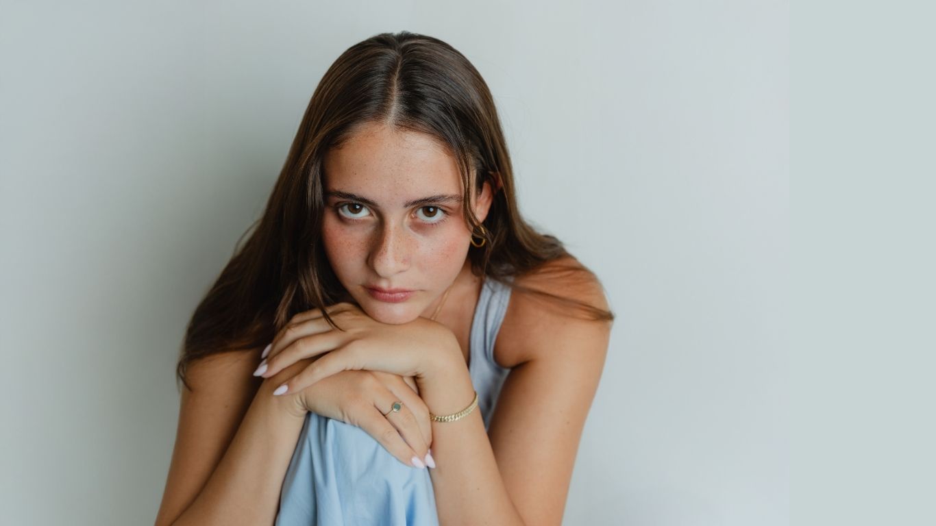 A young woman with long brown hair and light skin gazes intently at the camera, resting her chin and hands on her knee, wearing a light blue sleeveless top against a plain light background.