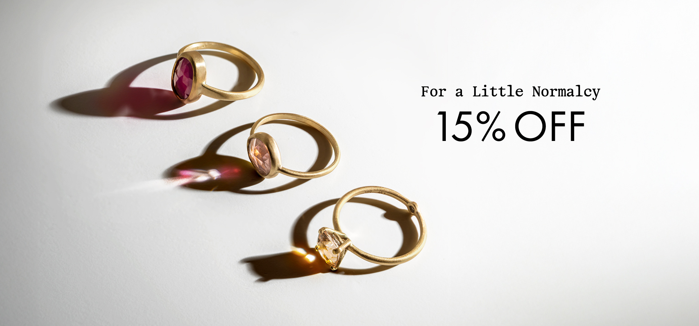 Three gold rings with colored gemstones are arranged on a white surface, casting soft shadows. Next to them, text reads For a Little Normalcy 15% OFF in black letters.